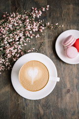 Delicious fresh morning cappuccino coffee with pink and red french macarons dessert on a plate on the rustic wooden table background with some blossoming flowers, top view