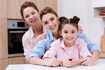 three generations of women standing in row together at kitchen and looking at camera