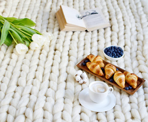 Cup with cappuccino and croissants, berries, white pastel giant knit blanket, bedroom, flowers tulips, spring, woman day, morning concept 