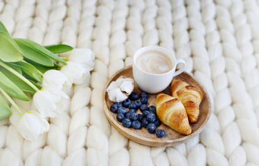 Cup with cappuccino and croissants, berries, white pastel giant knit blanket, bedroom, flowers tulips, spring, woman day, morning concept 