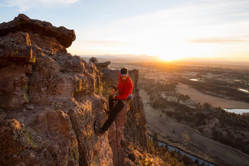 Obraz premium Adventurous man is rappeling down a cliff during a bright and vibrant sunny sunset. Taken in Smith Rock, Oregon, North America.