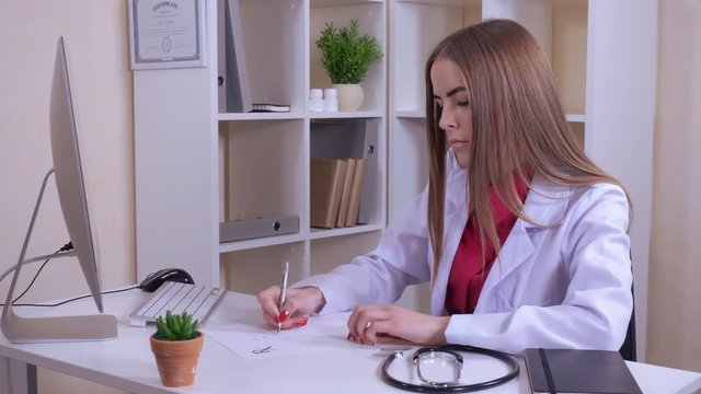 beautiful doctor sitting at the working place in own office writing prescription. professional caucasian therapist using internet on computer. female wearing uniform white coat stethoscope on the desk