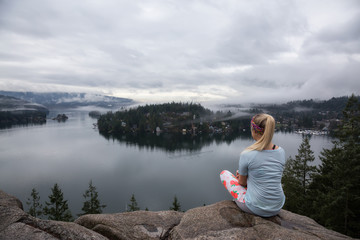 Young Caucasian Girl is enjoying a beautiful scenery from the top of a mountain during a cloudy winter morning. Taken in Quarry Rock, Deep Cove, North Vancouver, BC, Canada.