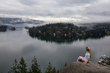 Young Caucasian Girl is enjoying a beautiful scenery from the top of a mountain during a cloudy winter morning. Taken in Quarry Rock, Deep Cove, North Vancouver, BC, Canada.