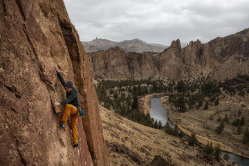 Adventurous man is rock climbing on the side of a steep cliff during a cloudy winter evening. Taken...