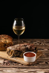 Pumpkin muffin and baked meat roll with filling, along with a sauce on a wooden plate and a glass of white wine on a black background