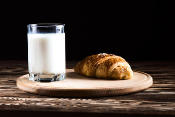 A delicious croissant on a round wooden plate near a glass of milk on a black background.