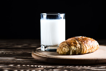 A delicious croissant on a round wooden plate near a glass of milk on a black background.