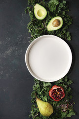 Green salad and fruits mix for salad or smoothie with kale, young beetroot leaves, sprouts, pear, avocado, pomegranate, white empty plate over dark texture surface. Top view, space. Food background.