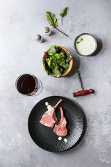 Grilled sliced rack of lamb with yogurt mint sauce served with green salad young beetroot leaves, glass of red wine, pink salt on round wooden slate board over grey texture background. Top view, space
