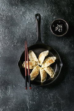 Asian dumplings Gyozas potstickers fried on cast-iron pan, served with chopsticks and bowl of soy sesame sauce over black texture background. Top view, space.