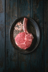 Raw uncooked black angus beef tomahawk steak on bone served with salt and pepper on vintage metal tray over dark wooden plank background. Top view, copy space.