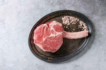 Raw uncooked black angus beef tomahawk steak on bone served with salt and pepper on vintage metal tray over grey texture background. Top view, copy space.