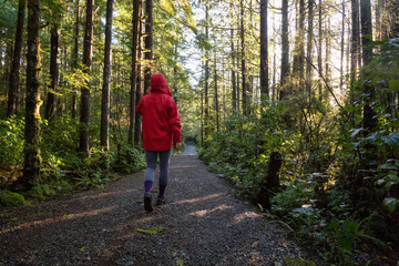 Obraz premium Girl wearing a bright red jacket is walking the the beautiful woods during a vibrant winter morning. Taken in Ucluelet, Vancouver Island, BC, Canada.