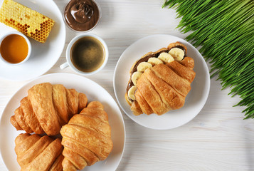 Croissants with chocolate butter and banana, coffee, honey and sprouted wheat on a wooden background. Breakfast flat lay.