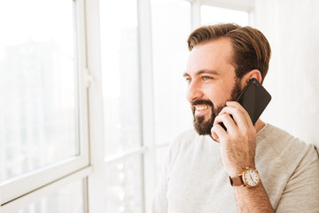 Closeup portrait of cheerful guy with beard and mustache looking through window, while having pleasant mobile call