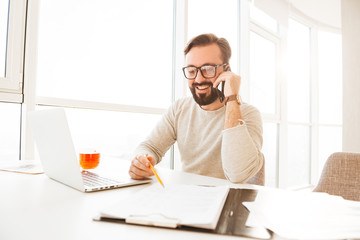 Businesslike man 30s in casual clothing smiling and negotiating about work using cell phone, while...