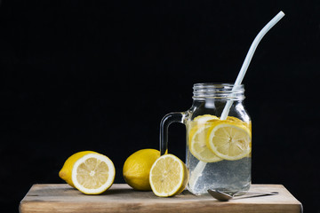 Close up of lemonade in glass jar with straw and fresh sliced lemons on wooden board with black background