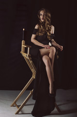 An attractive stylish young woman in a long black dress sits on a chair in a dark room. Studio photo. Fasche photo.