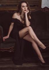 An attractive young woman in a long black dress with a deep neckline, with long legs sitting on the couch. Studio photo. Fashion photo.