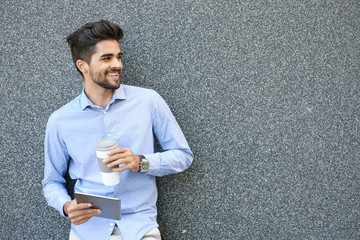 Young businessman leaning against wall with digital tablet. Man holding cup of coffee.