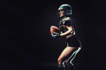 Poster Sportive serious woman in helmet of rugby player holding ball in stuio on dark background. © zamuruev