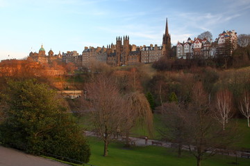 Widok na zabudowę Edynburga, Szkocja, z parku, na pierwszym planie drzewa i zieleń parku, w tle, na wzgórzu historyczne budynki, wieża kościoła, kamienice, piękne złote światło zachodzącego słońca