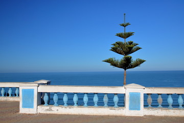 Piękny widok na morze, na pierwszym planie biało-niebieska ozdobna balustrada, po prawo nieduże zielone drzewko, balustrada w style andaluzyjskiej architektury, błękitne niebo - obrazy, fototapety, plakaty