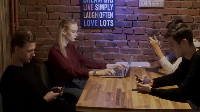 Four students are sitting at the wooden table in cozy cafe and surfing the internet. Blond woman with bordo lips is tping messege on her brand nwe laptop and three men in white and black jumpers are