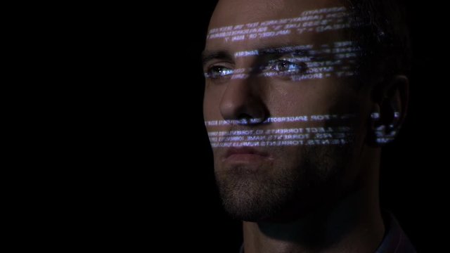 Close up of an adult male IT programmer working in a dark office room while the sequential code is reflected on his face