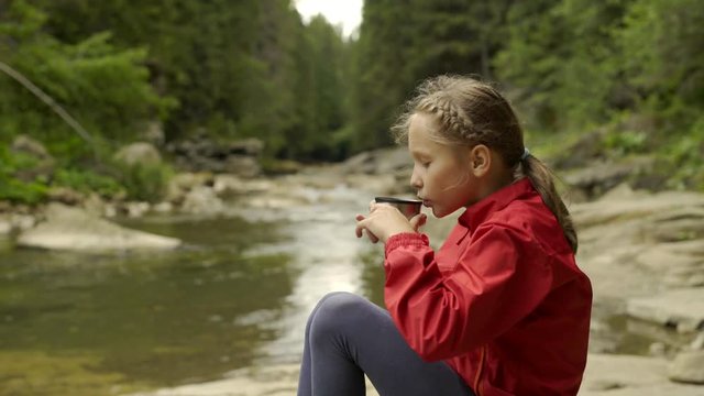 Sliding video of a child sitting on the bank of mountain river with cascades in forest drinking hot drink