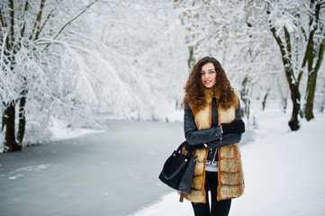 Elegance curly girl in fur coat at snowy forest park at winter.