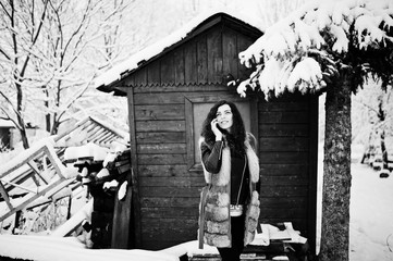 Fototapeta na wymiar Elegance curly girl in fur coat at snowy forest park agasinst green wooden house at winter.
