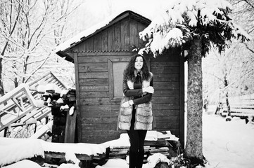 Elegance curly girl in fur coat at snowy forest park agasinst green wooden house at winter.