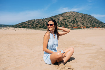 Brunette in sky blue dress sits and looks away in lotus pose on the sand and listens music in the earphones. Green mountain on background