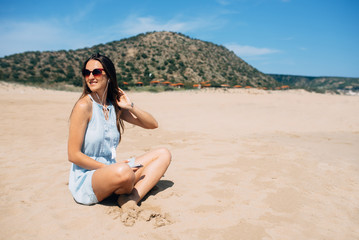 Brunette in sky blue dress sits and looks away in lotus pose on the sand and listens music in the earphones. Green mountain on background