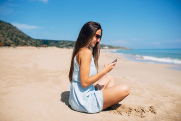 Fototapeta na wymiar Smiling girl in sky blue dress with sunglass on her face sits in lotus pose on the beach and looks in her cell phone. Sea waves on background