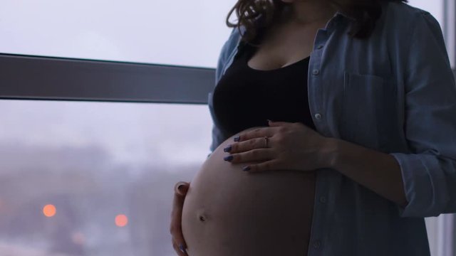 A happy pregnant woman stands at a large panoramic window located at a high altitude. He touches his stomach with his hands and communicates smilingly with the baby in the stomach. She looks at the