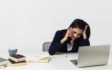 The business woman is working at office hour ,plenty of paper work and books stacked on table,she is using mobile phone and looking at laptop for check the data,busy work.