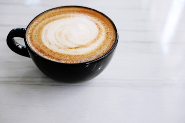 Hot coffee cappuccino latte in black cup  with stirred spiral milk foam on white marble texture table background, close up.