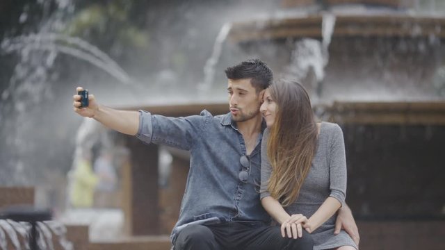 Happy couple pull faces for selfies together in front of a water fountain