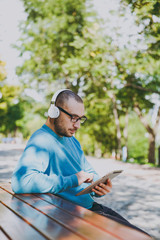 Obraz premium Young happy man, businessman or student in casual blue shirt glasses sitting at table with headphones, tablet pc in city park, listen music, rest outdoors on green nature. Lifestyle leisure concept.