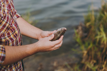 Close up Man in checkered shirt with rolled up sleeves caught a fish and holds it in arms on the shore of lake on background of water, shrubs and reeds. Lifestyle recreation, fisherman leisure concept