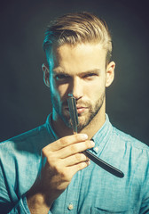 Bearded handsome, sexy macho with straight razor in hands poses in studio. Young hairdresser wearing stylish shirt demonstrating sharp blade of his dangerous razor.