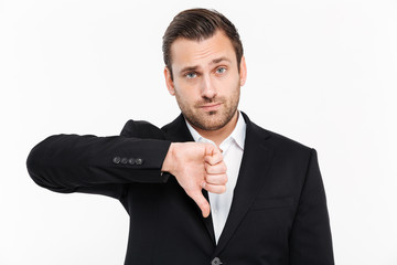 Portrait of displeased male office worker wearing black formal suit and showing thumb down on camera with disappointment, isolated over white wall