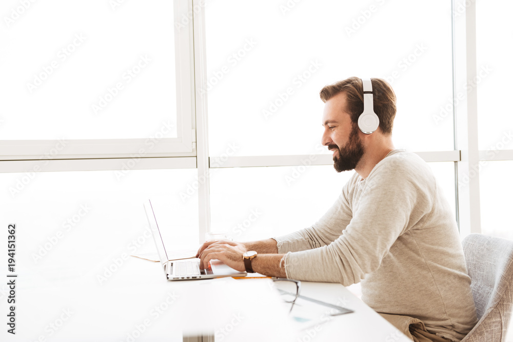 Sticker Image in profile of modern guy 30s using white headphones and communicating on laptop, while resting in hotel apartment