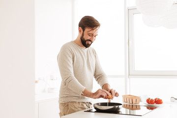 Image of cheerful single man wearing casual clothing cooking omelet with tomato in kitchen, while having breakfast in modern flat