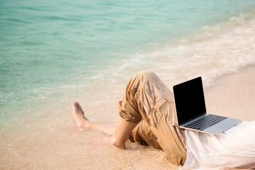 Asian man with his computer on the beach with waves from sea and golden sunlight at evening. working on computer at outdoors while traveling at sea in summer holiday. freelancer work concept.