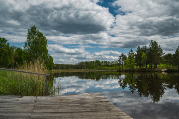 Lake in the green forest. Forest lake. Beautiful summer landscape with forest, lake. Cloudy sky reflected in water. Wooden pontoon on a forest lake