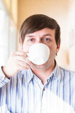 A white middle-aged man drinks tea or coffee from a white cup, bright sunlight. Look straight at the viewer. Morning or day.